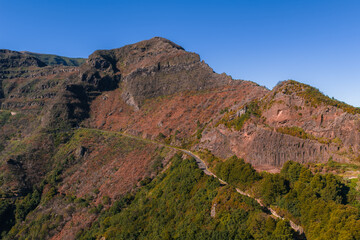 Obraz premium Aerial view of rugged volcanic ridges and terraced slopes on Madeira, Portugal, near Pico do Arieiro. A narrow path winds mid slope in clear midday light.