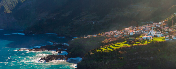 Whitewashed houses with terracotta roofs sit on steep Madeira cliffs, green terraces border black volcanic rocks and foaming surf in late afternoon high contrast light. © Aerial Film Studio