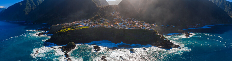 Aerial panorama shows a cliff ringed peninsula on Madeira Island, Portugal, whitewashed houses on lava rock, sea stacks, surf on basalt, roads and terraced fields in low light. © Aerial Film Studio