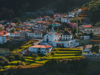 Whitewashed houses with terracotta roofs surround a church with a teal bell tower in Madeira, Portugal, as terraced fields step toward a coastal cliff in late afternoon light. © Aerial Film Studio