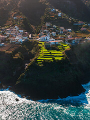 Terraced fields and whitewashed houses descend a cliff above Atlantic waves in Madeira, Portugal, near Santana or Sao Vicente, in warm late afternoon light. © Aerial Film Studio