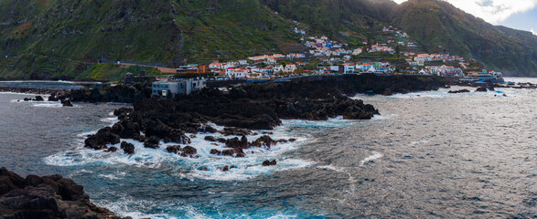 Rugged volcanic shore meets Atlantic surf at Porto Moniz, Madeira. Black basalt pools sit by a compact hillside town. Cloudy late afternoon light adds a moody tone. © Aerial Film Studio