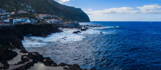 Rugged black lava pools meet deep blue Atlantic at Porto Moniz, Madeira. White houses line steep green cliffs under bright daylight with scattered clouds. © Aerial Film Studio