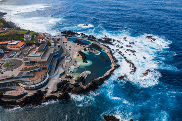 Aerial view shows seawater pools in black lava at Porto Moniz, Madeira, with walkways, terraces, and facilities. Turquoise basins meet blue surf under bright daylight.