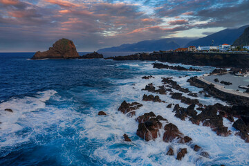 Aerial view shows Porto Moniz tidal pools, Ilheu Mole, and village on Madeira, Portugal. Waves hit black basalt, pastel sunset clouds glow, wide angle shows motion and form. © Aerial Film Studio