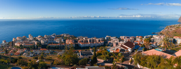 Panoramic view of Funchal, Madeira, with terracotta roofs, white buildings, and palm streets. Daylight reveals hotels, terraces, gardens, and a headland by the sea. © Aerial Film Studio