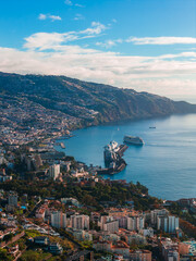Elevated daylight view of Funchal, Madeira Island, Portugal. Two cruise ships dock at the Port of Funchal pier, with terraced hills, red roofs, and a calm deep blue bay. © Aerial Film Studio
