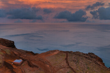 Reddish brown cliffs descend to calm Atlantic water at dusk in Madeira, Portugal. A small metal roof hut sits on a barren plateau, with a faint trail on the headland.