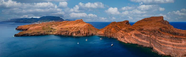 Fototapeta premium Aerial view shows rust red cliffs of Ponta de Sao Lourenco, Madeira, Portugal, small sailboats in a calm bay, bright midday light, cumulus clouds, cobalt Atlantic