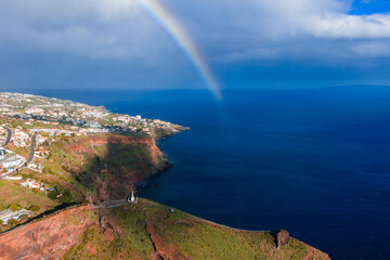 Rugged red and green cliffs meet the Atlantic near Garajau, Madeira. A faint rainbow arcs as sunlight breaks through showers over white homes and terraces toward Funchal. © Aerial Film Studio