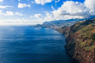 Fototapeta premium Aerial view shows Madeira south coast near Cabo Girao, reddish cliffs meet Atlantic, Funchal spreads along shore, layered mountains rise, soft light and clouds present.