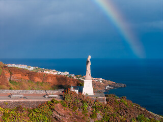 Cristo Rei at Garajau stands on a pedestal above the Atlantic in Madeira, Portugal. Terraced roads, coastal scrub, and white houses appear as a rainbow arcs under post storm light. © Aerial Film Studio