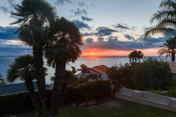 Sunset light warms palm silhouettes, hedged terraces, and red roofs near Funchal, Madeira. Orange clouds glow over the Atlantic as wide framing presents the ocean horizon. © Aerial Film Studio