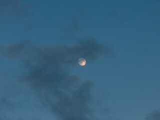 A pale near full moon sits in a clear blue evening sky as soft wispy clouds drift by. No horizon is visible. Gentle twilight light reveals faint crater detail.