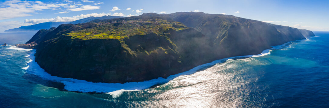 Aerial panorama of Madeira Island headland in Portugal, volcanic cliffs meet deep blue water, white surf wraps the base, terraced fields and plateaus glow in clear daylight