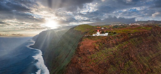 Aerial view shows white lighthouse on rust red cliffs at Ponta do Pargo, Madeira, Portugal, sunbeams through clouds, waves below, villages on plateau, near sunset.