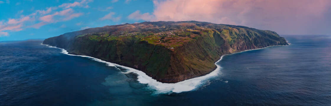 Aerial panorama shows a triangular headland on Madeira, Portugal, with cliffs, foaming Atlantic surf, fields, villages, and roads in warm late day light and pastel tones.