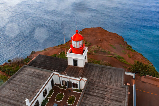 Aerial view shows Ponta do Pargo Lighthouse on Madeira, Portugal, set on rugged cliffs above the Atlantic, with calm seas, overcast light, and a geometric courtyard.