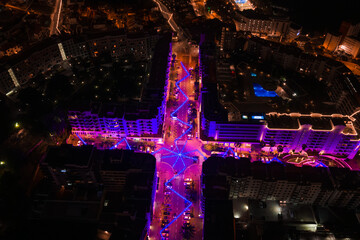 Aerial night view of a Funchal, Madeira intersection at night, neon purple and magenta lights form zigzags and star nodes, hotel avenues and a lit rooftop pool on right.