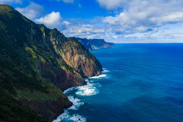 Aerial view of volcanic cliffs on Madeira north coast, Portugal, waves strike dark rock and green streaks, headlands recede, sunlit patches cut through scattered clouds.