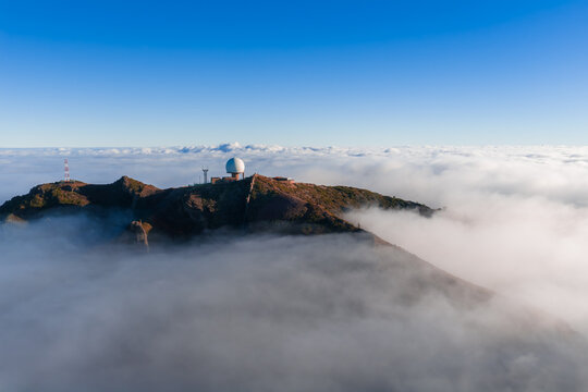 Aerial view shows Pico do Arieiro on Madeira, Portugal, with a white radar dome and antennas above clouds, reddish rock, and wide framing in clear morning light.