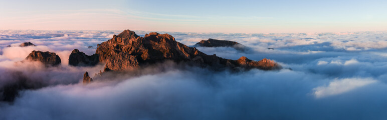 Aerial view of jagged volcanic peaks near Pico do Arieiro, Madeira, Portugal, rising above clouds at golden hour, warm light on rock faces and pastel sky.