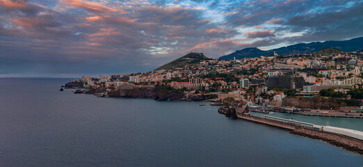 Panoramic coastal view of Funchal, Madeira, Portugal, with harbor, breakwater, marina, volcanic headlands, and white terracotta buildings rising on green hills. © Aerial Film Studio