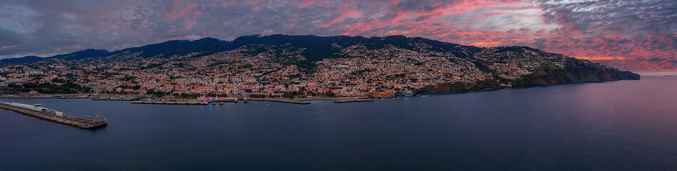 Wide panoramic view of Funchal, Madeira, at dusk. Harbor breakwater extends into dark blue Atlantic waters, white buildings climb steep slopes, city lights glow.
