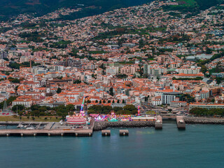 Aerial view of Funchal, Madeira, Portugal at dusk shows hillsides, terracotta roofs, waterfront promenade, marina, and a lit fairground by the harbor with calm waters.