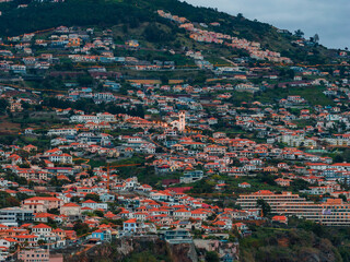 Funchal, Madeira, Portugal hillside town at dusk shows white houses, terracotta roofs, winding lit roads, and a tall church bell tower across steep layered slopes. © Aerial Film Studio