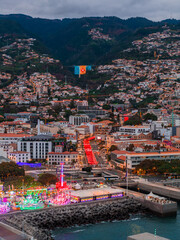 Aerial view of Funchal, Madeira at dusk shows marina and fairground, a giant illuminated Portuguese flag on a valley slope, red holiday lights along an avenue, and terraced homes. © Aerial Film Studio