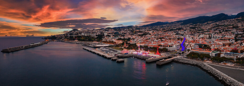 A panoramic aerial view shows Funchal, Madeira at dusk, terraced hills and white roofs leading to the marina, yachts and breakwater, fairground and purple lit CR7 Museum.