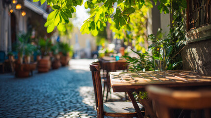 Fototapeta premium Sunlit outdoor caf? seating with wooden tables and chairs shaded by lush green foliage on a charming cobblestone street in a peaceful urban setting
