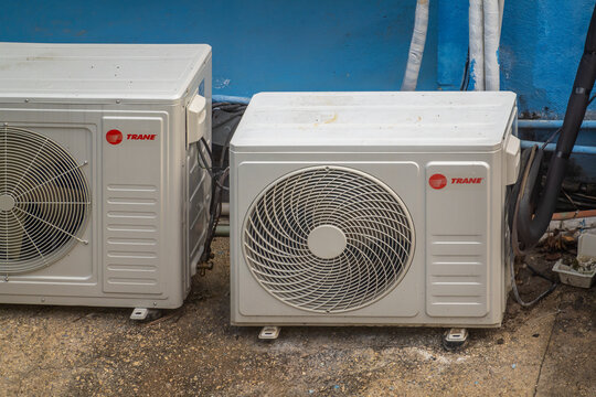 Oaxaca, Mexico. Dec 12, 2025. A pair of white, branded Trane air conditioning condenser units sit on a concrete floor next to a vivid blue exterior wall with exposed wiring.