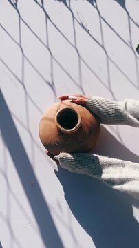 a pair of hands gently holding a weathered clay vessel under natural light with soft geometric shadows.