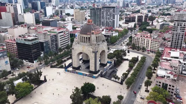 Aerial panoramic view of Plaza de la Rep&uacute;blica as the drone orbits the Monument to the Revolution, revealing the surrounding city blocks while people walk across the open square.