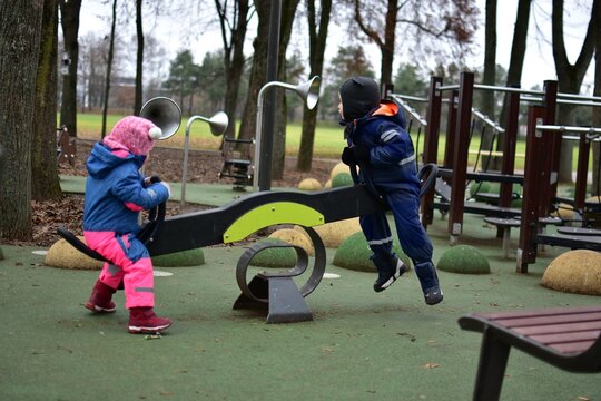 Two toddlers playing together on playground seesaw in autumn park. Children in warm winter outfits enjoy cooperative outdoor play, learning balance and social skills.