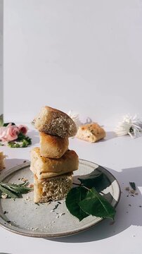 a small stack of golden focaccia bread pieces resting on a ceramic plate, surrounded by herbs, leaves, and soft floral accents on a clean white surface.