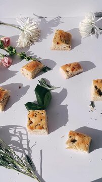 Softly arranged rosemary focaccia bread cubes scattered across a white surface with fresh herbs and pale florals, creating a calm and organic