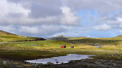 Traditional red cabins in remote mountain landscape featuring rolling green hills, dramatic clouds and a small pond. Vestland, Norway, travel Europe