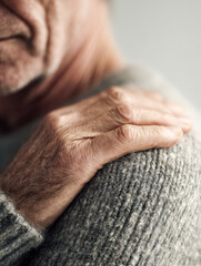 Fototapeta premium Elderly person experiencing shoulder discomfort gently holding the affected area while wearing a cozy knitted gray sweater indoors with soft lighting