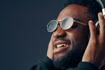 Happy young man enjoying music with headphones, wearing stylish sunglasses against a dark...