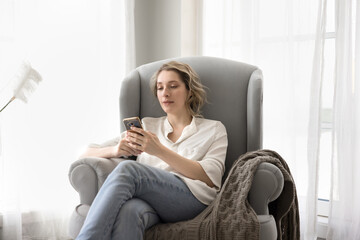 Cozy chatting. Calm beautiful mid 30s woman unwind at home sitting in soft armchair typing message...