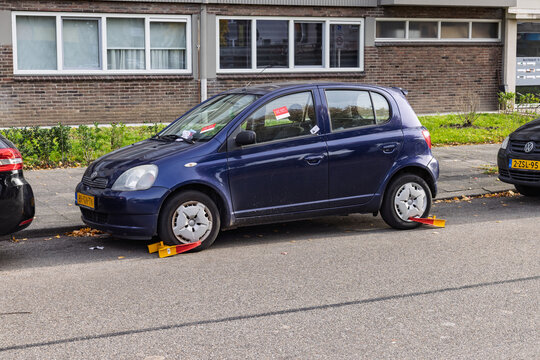 A parked blue car immobilized with two yellow and red wheel clamps on the tires due to a parking violation in a Dutch urban street setting.