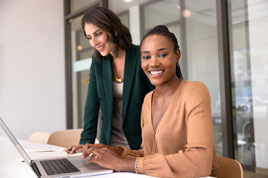 Happy young African American professional woman working at computer with female colleague, looking at camera with toothy smile. Portrait of multiethnic intern and mentor