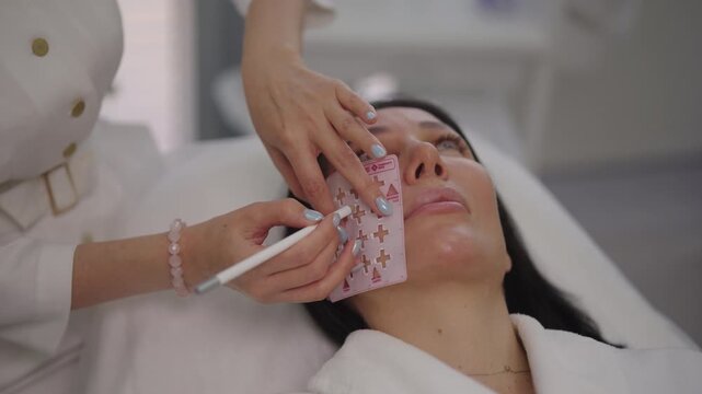 Close-up hands of professional beautician preparing female client for facial cosmetic procedure, using skin marking pencil and stencil to outline treatment areas for enhanced beauty, slow motion.