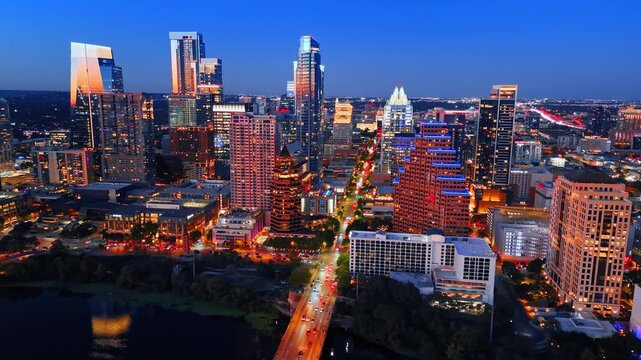Transport with numerous cars with headlights on cross the bridge above the Colorado River. Amazing scenery of Austin downtown, Texas, USA with beautiful illumination. Aerial view.