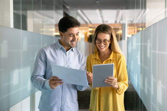 Happy young coworkers comparing sales reports, analyzing paper document and electronic content on tablet, standing in office hall, corridor, talking, discussing financial analytic data