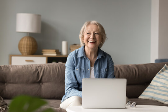 Enjoying digital victories. Happy senior woman active retiree in stylish clothes spend leisure time at home office by notebook look aside with successful smile proud of confident virtual achievements