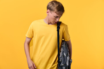 Boy in a yellow t shirt with a camouflage backpack stands against a bright yellow backdrop, casual...